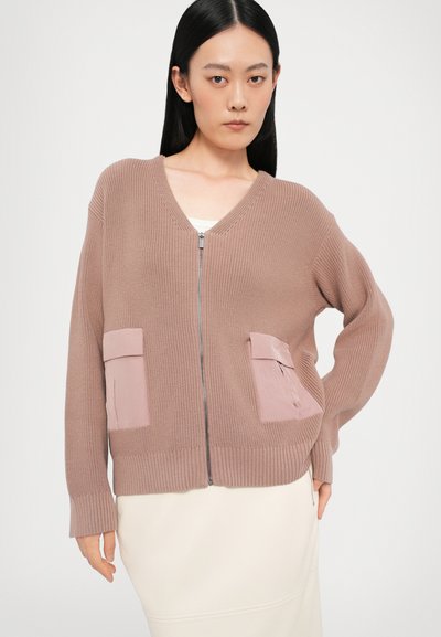 Garo griezuma adījuma krekliņš gaiši brūnā krāsā ar V veida kakla izgriezumu, kuram ir divas priekšējās kabatas kontrastējošā gaiši rozā materiālā un ribu apkakles.