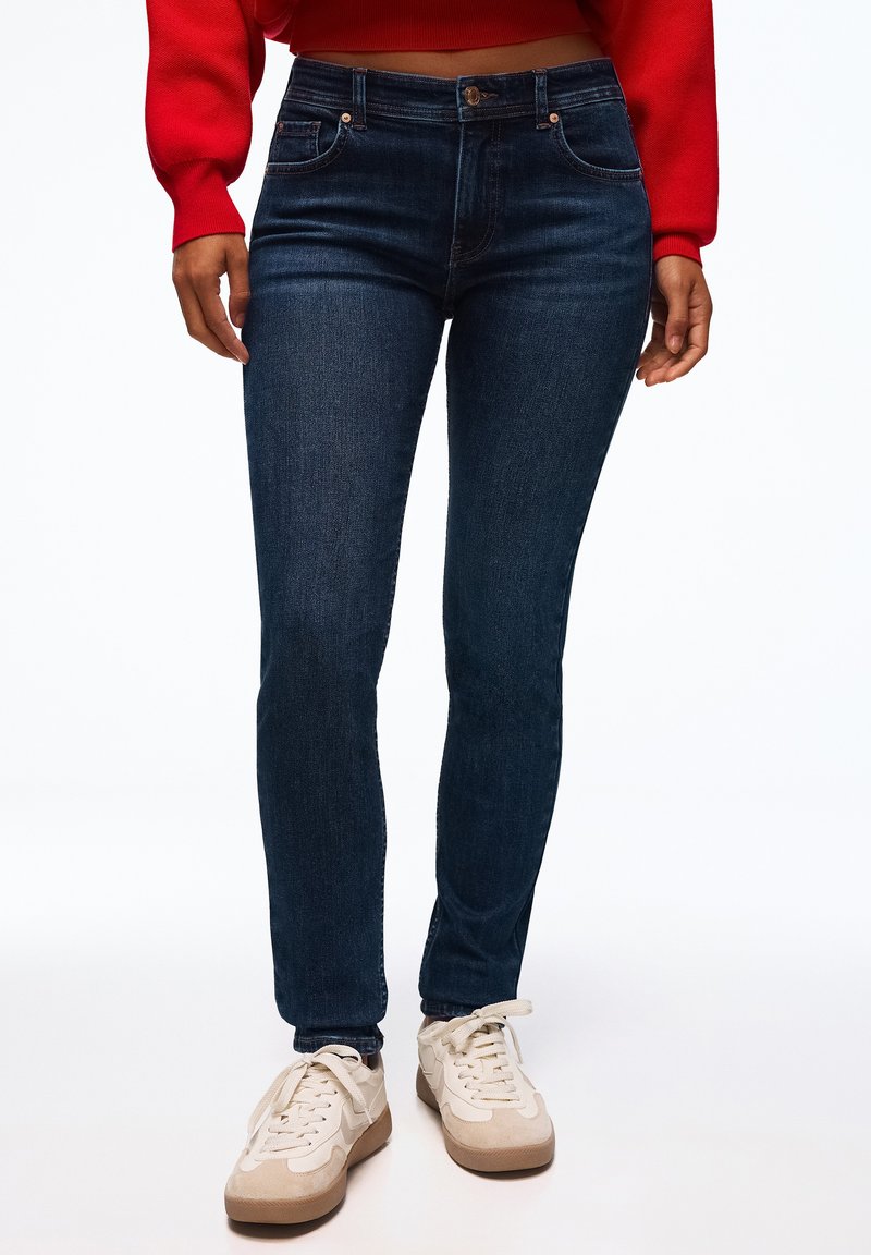 Personne portant un jean skinny bleu foncé, des baskets beige et blanches, et un haut rouge court à manches longues, debout devant un fond blanc.