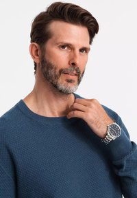 Uomo che indossa un maglione blu scuro a trama con collo girocollo, con un orologio argentato dal quadrante nero e cinturino in metallo.