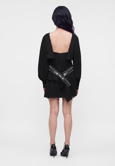 Femme aux longs cheveux foncés porte une robe noire avec un dos largement ouvert, ceinture cloutée, manches longues et talons noirs, debout de dos.