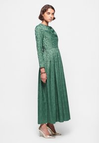 Grøn lang kjole med blomsterprint, cowl-udskæring, tætsiddende bodice og plisseret nederdel, parret med metallic guld højhælede sko.
