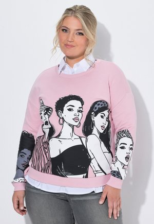Blonde Frau trägt einen pinkfarbenen Sweatshirt mit illustrierten vielfältigen weiblichen Gesichtern, geschichtet über einem weißen Hemd mit Kragen und grauer Jeans.