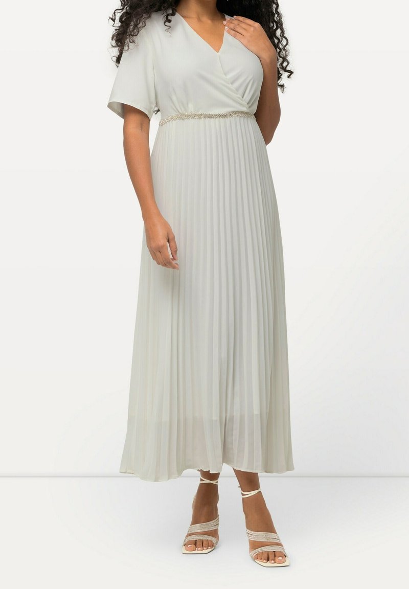 Femme portant une robe maxi plissée gris clair à manches courtes avec une ceinture décorative, associée à des sandales à talons hauts et à brides.