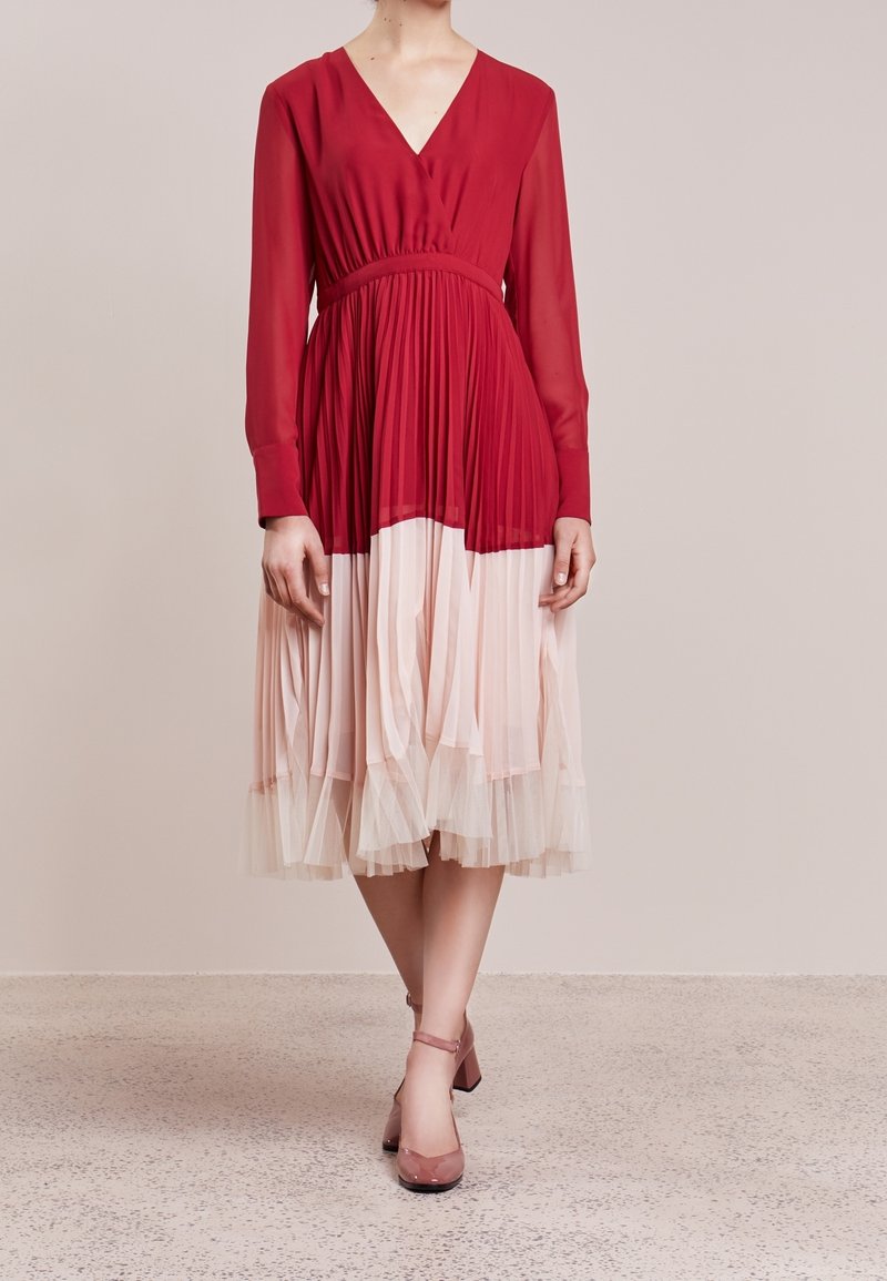 Rotes, plissiertes Kleid mit langen Ärmeln, einem V-Ausschnitt und einer taillierten Taille. Der Rock verblasst von Rot zu hellrosa und ist mit transparenter Stoffschicht versehen. Rosa Schuhe.