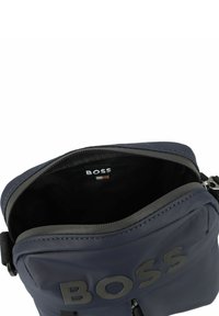 Námorně modrá nylonová crossbody taška s texturovaným povrchem. Obsahuje výrazné černé logo, zipový uzávěr a nastavitelný popruh. Interiér je tmavý a hladký.