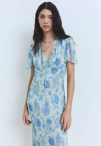 Hellblaues Blumenkleid aus fließendem Stoff. Mit kurzen Ärmeln, V-Ausschnitt, Zugbanddetail und figurbetonender Silhouette. Goldene Halskette.