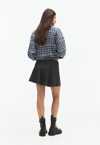 Chemise oversize à carreaux bleus, jupe évasée noire et bottes en cuir noir à lacets montantes. Fond minimaliste.