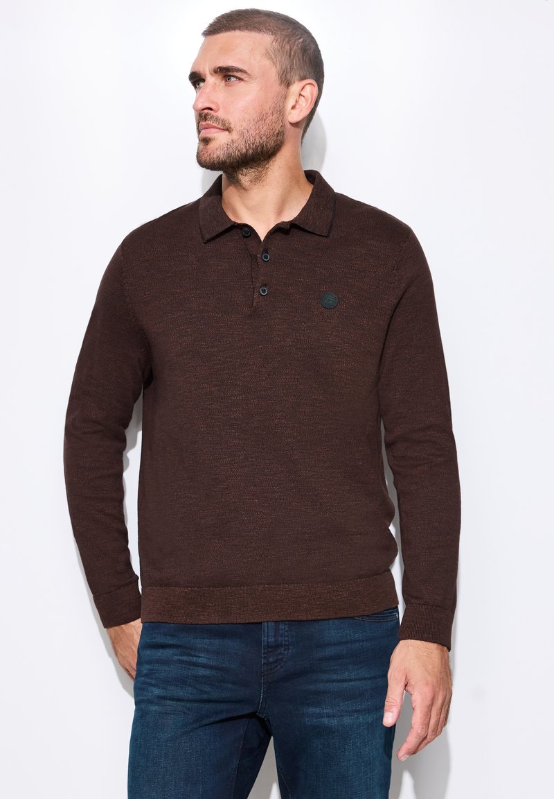 Homme portant un polo à manches longues marron foncé et un jean bleu, regardant vers sa gauche sur un fond uni.