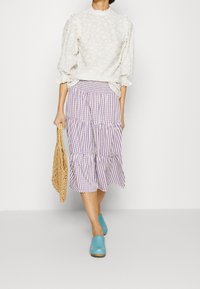 Blouse blanche brodée à manches bouffantes, associée à une jupe à volants à carreaux violets et blancs, et des mules bleues, tenant un sac en osier beige.