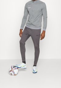 Grijze sportjas met rits, gecombineerd met grijze slim-fit broek, met een subtiel logo. Witte en blauwe sportschoenen, met een meerkleurige voetbal.