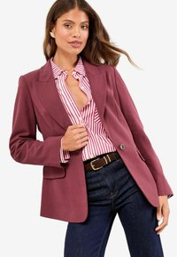 Bordeaux blazer met een getailleerd ontwerp, met klepjes en twee voorzakken, gedragen over een rood-wit gestreept shirt. Donkerblauwe jeans.