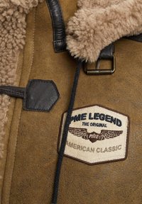 Bruin schapenvachtjack met een beige fleece-voering bij de kraag, leren accenten en een opvallende crèmekleurige en marineblauwe merkpatch met vleugel-logo.