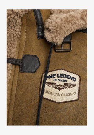 Smeđi jahalni kaput od ovčije kože s bež krznom koja oblaže ovratnik, kožnim detaljima i istaknutim kremastim i mornarsko plavim logotipom brenda s krilima.