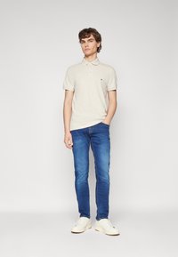 Polo shirt beige chiaro con maniche corte e colletto, abbinato a jeans slim-fit blu scuro e sneakers bianche. Nessun logo visibile sui pantaloni.
