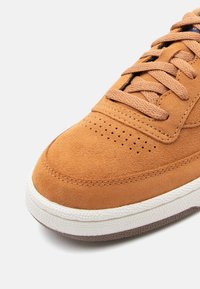 Adidași din suede maro cu un superior texturat, având perforații, șireturi plate și o talpă din cauciuc albă cu un accent gri.