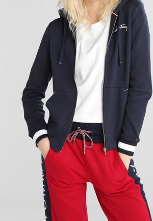 Personne portant un sweat à capuche zippé bleu marine par-dessus une chemise blanche et un pantalon rouge à cordon avec des bandes latérales bleu marine, les mains dans les poches.