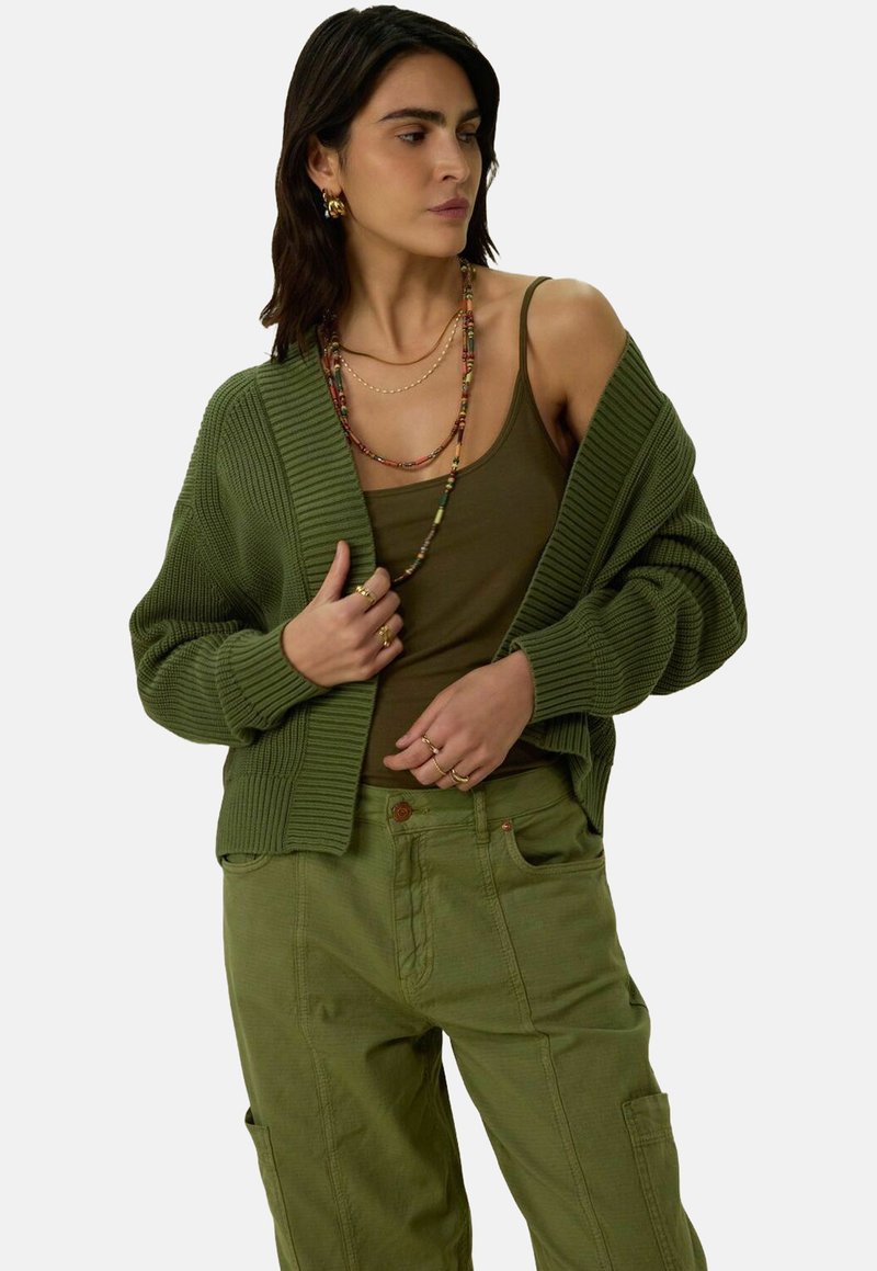 Cardigan en tricot vert olive avec une coupe décontractée et une texture côtelée, associé à un débardeur vert et un pantalon assorti. Colières en couches incluses.