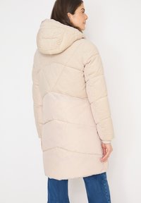 Kvindelig iført en beige quiltet puffjakke med corduroy på den øverste ryg og hætte, stående sidelæns med afslappede hænder, blå jeans synlige.
