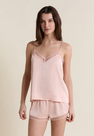 Junge Frau mit braunen Haaren, die ein zartrosa Satin-Top und Shorts mit Spitzensaum trägt, steht vor einem schlichten hellen Hintergrund.