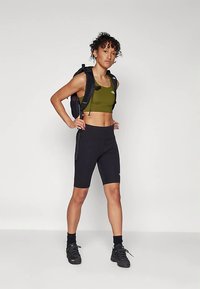 Pantaloncini da ciclismo neri abbinati a un reggiseno sportivo verde oliva, indossati sotto uno zaino nero. L'outfit presenta un design aderente e atletico.