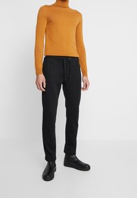 Pantalones negros de corte ajustado con cordón y bolsillos, combinados con un suéter de cuello alto ajustado en un cálido color mostaza. Material liso y líneas limpias.