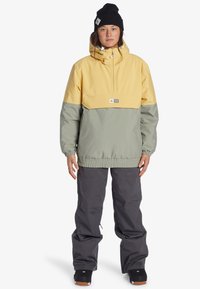 Pull-over jacka i gult och grönt, med hög krage, justerbar dragsko på huvan och elastiska ärmslut. Matchad med grå byxor.
