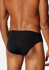 Slip da bagno neri, tessuto liscio, design a vita media, copertura minima, cinturino elastico, accentua le forme del corpo, senza motivi o accessori visibili.