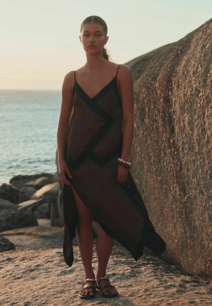 Frau trägt ein dunkles, durchsichtiges Kleid mit schwarzem Rand und Sandalen, steht an einem felsigen Ufer in der Nähe eines großen Felsens und des Ozeans bei Sonnenuntergang.