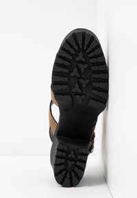 Chaussure marron et noire avec semelle en caoutchouc texturée, présentant des crampons profonds et des sangles. Semble durable avec un design de panneaux distincts. Taille marquée 37.