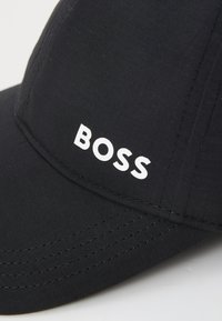 Black cap made of textured fabric featuring a curved brim and stitched detailing. The brand name "BOSS" is prominently displayed in white.