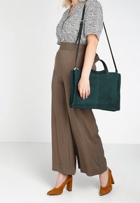 Nainen, jolla on päällään kuviollinen paita ja ruskeat leveälahkeiset housut, kantaa tummanvihreää mokkalahjakassia ja jalassaan on ruskeat mokkasaappaat.