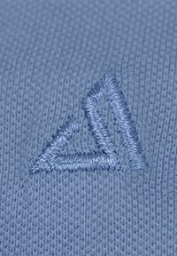Hellblaue gestrickte Stoff mit einem detailliert gestickten, dreieckigen Logo in einem etwas dunkleren Blauton. Strukturiert und eng gewebt.