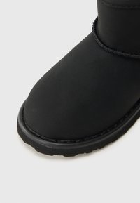 Close-up van een zwarte winterlaars met matte afwerking, ronde neus, dikke zool en gevlochten randdetail nabij de enkel.