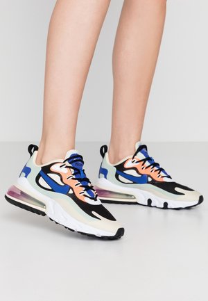 Personne portant des baskets de sport multicolores avec des accents bleus, orange, noirs et blancs, se tenant sur un sol gris clair uni.