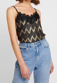 Camisole s černými a zlatými zigzag sequiny s jemným černým krajkovým lemováním, s tenkými nastavitelnými ramínky, spárovaný s vysokými světle modrými džíny.