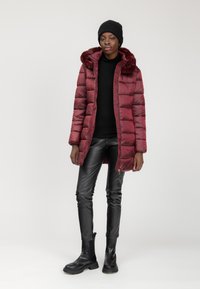 Mujer de pie con un gorro negro, chaqueta acolchada burdeos con cuello de piel, suéter negro de cuello alto, pantalones de cuero negros y botas negras sobre un fondo blanco.
