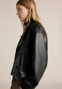 Veste en cuir noir courte avec un large col, deux poches avant et des détails en boutons à l'avant et aux manches. Texture lisse.