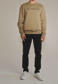 Felpa khaki con la scritta "diverse" in un font più scuro, abbinata a pantaloni neri e sneaker bianche, con scollo a girocollo e polsini a costine.