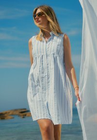 Blauw-wit gestreepte jurk met knoopsluiting aan de voorkant en een losse pasvorm, gedragen met een zonnebril tegen een strandachtergrond.