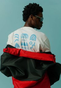 Bílá tričko s modrými grafickými vzory na zadní straně, vrstvené pod černou a červenou bundou, nošené s slunečními brýlemi na světle modrém pozadí.