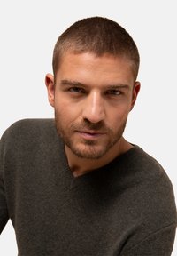 Tmavě šedý svetr s výstřihem do V, měkká textura, žebrovaný límec, nošený modelem s krátkými vlasy a vousy, na prostém bílém pozadí.