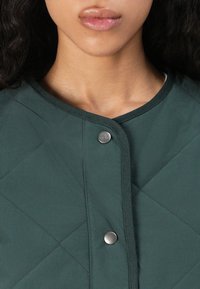 Veste verte matelassée avec un col rond, fermeture à boutons-pression et texture lisse ornée de coutures ton sur ton.