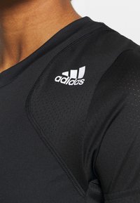 Musta urheilupaita, jossa on pyöreä kaula-aukko, tekstuuriset hartiapaneelit ja valkoinen Adidas-logo vasemmassa yläkulmassa. Suora, istuva muotoilu.