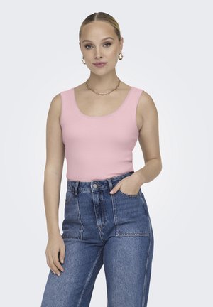 Donna in piedi con una mano in tasca, indossa un top rosa senza maniche, jeans blu a vita alta, orecchini a cerchio dorati e una collana a catena.