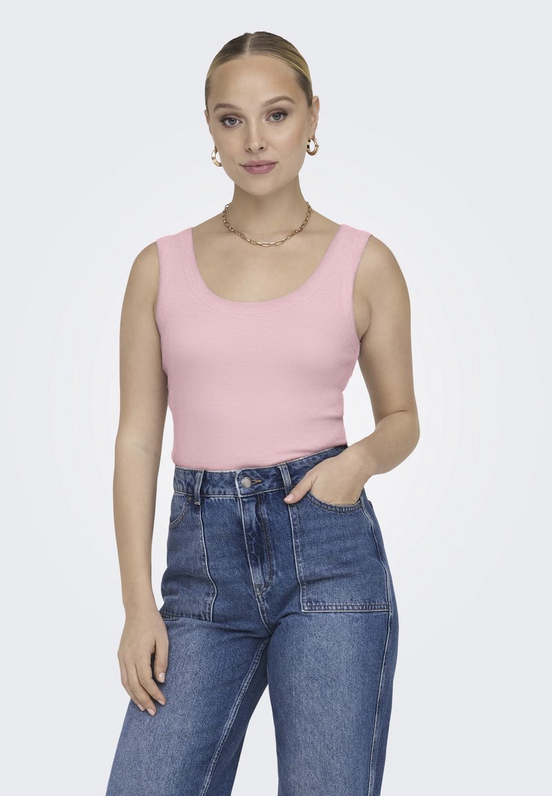 Donna in piedi con una mano in tasca, indossa un top rosa senza maniche, jeans blu a vita alta, orecchini a cerchio dorati e una collana a catena.