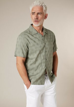 Middelbare man met grijs haar, gekleed in een olijfgroen overhemd met korte mouwen met patroon en witte broek, handen in de zakken, kijkend naar de zijkant.