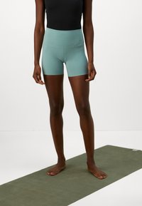 Persoon die blootsvoets op een groene yogamat staat, met lichtteal hoge sportshorts en een zwart mouwloos shirt.