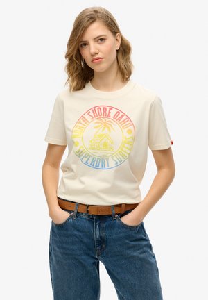 T-shirt en coton crème ornée d'un graphique circulaire coloré avec des palmiers et du texte. Associée à un jean en denim bleu taille haute et une ceinture marron.