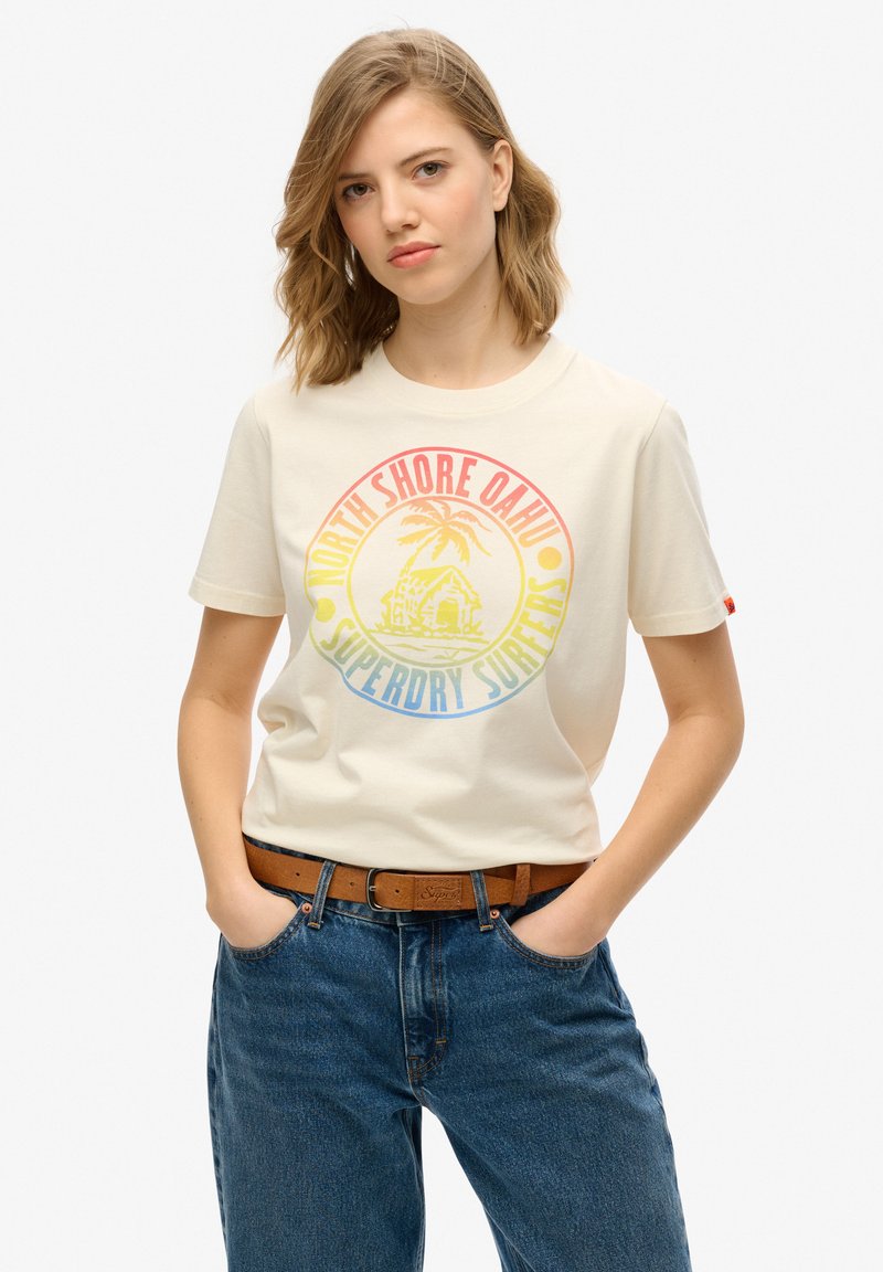 Camiseta de algodón color crema con un gráfico circular colorido que presenta palmeras y texto. Combinada con unos vaqueros azules de tiro alto y un cinturón marrón.