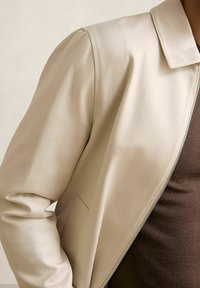 Beige lynlåsjakke med en glat overflade, en subtil glans, en nedslået krave og en sidelomme. Bæres over en brun skjorte.